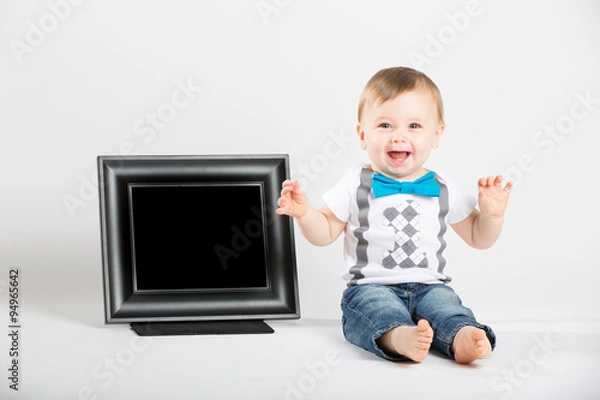 Fototapeta a cute 1 year old baby sits next to a blank black picture frame in a white studio setting. The boy is extremely excited with hands in the air. dressed in Tshirt, jeans, suspenders and blue bow tie