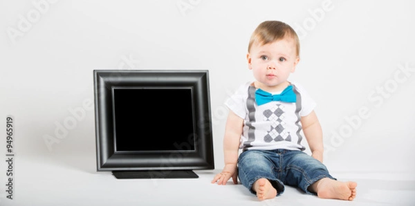 Fototapeta a cute 1 year old baby sits next to a blank black picture frame in a white studio setting. The boy has a confused expression. He is dressed in Tshirt, jeans, suspenders and blue bow tie