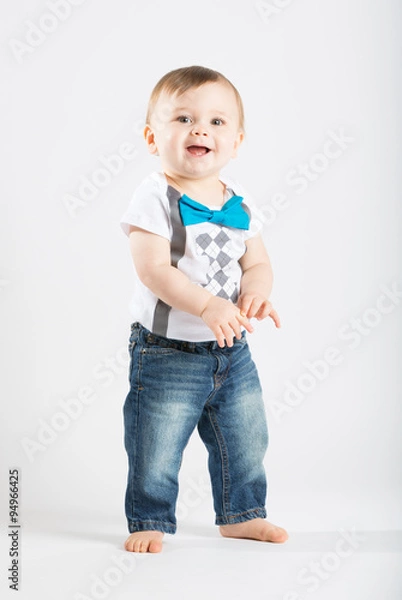 Fototapeta a cute 1 year old stands in a white studio setting. The boy has a happy expression. He is dressed in Tshirt, jeans, suspenders and blue bow tie