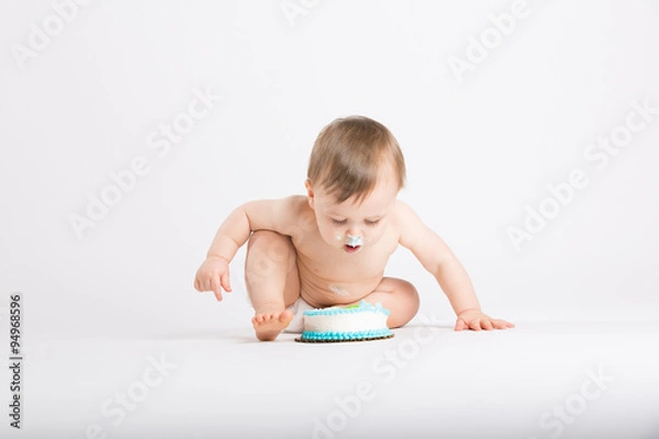 Fototapeta a cute 1 year old sits in a white studio setting. The boy in the middle of the shot about to get another face full of cake. He is only dressed in a white diaper