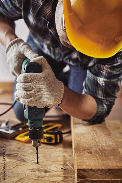 Fototapeta Young handyman in hardhat drilling wood in working studio. Concept of craft and technology.