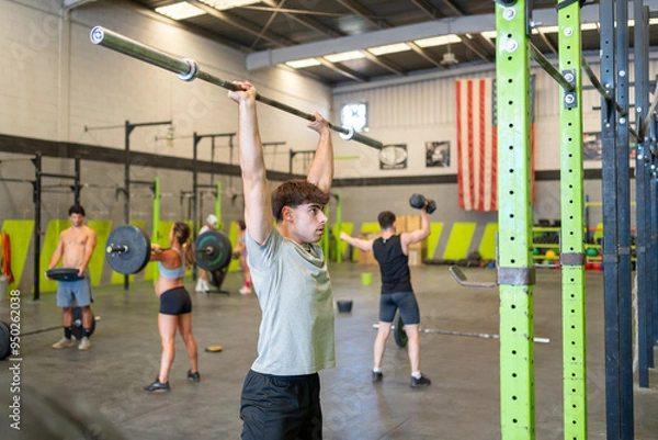 Obraz A guy warming up with a barbell in the gym before doing crossfit with a group of people training behind him.