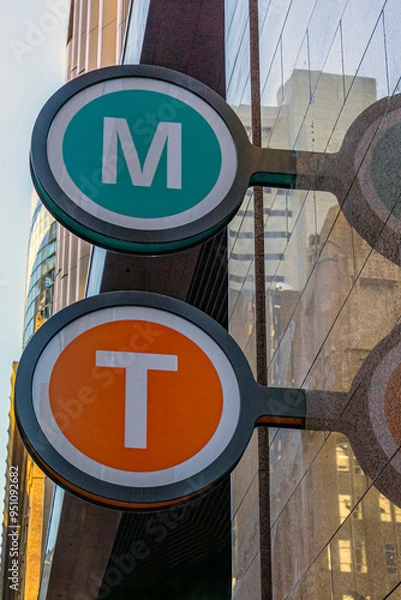 Fototapeta Close-up of Train and Metro Sign, in Sydney, Australia. Round Green and Orange Signs with Letters T and M, attached to the entrance wall to a Train and Metro Station. Photo taken in July, 2024.