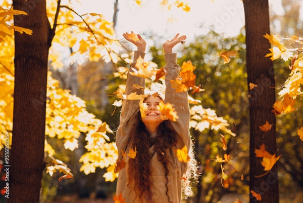 Fototapeta Smiling young woman with hands up throws up the autumn leaves in the forest at sunset