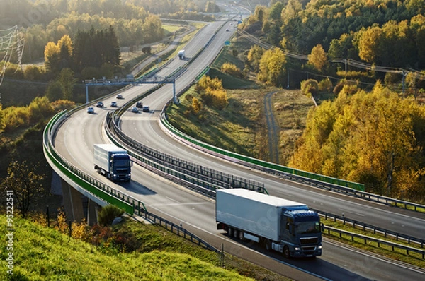 Fototapeta Asfaltowa autostrada z elektronicznymi bramkami poboru opłat w jesiennym lesie. Trzy ciężarówki na drogach. Most łączący dolinę. Widok z góry. Słoneczny dzień w jasnych kolorach jesieni.
