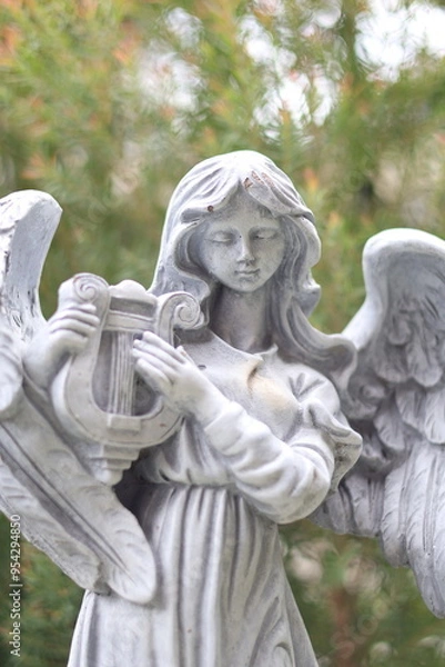 Obraz A close-up photo of a white stone statue depicting an angel with wings, playing a harp. The angel has a serene expression and is surrounded by lush greenery.