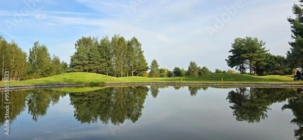 Obraz A golf green with a white flag near a pond, reflecting trees and the sky in the water. Peaceful scene with natural colors.