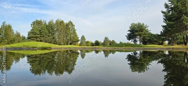Obraz A golf green with a white flag near a pond, reflecting trees and the sky in the water. Peaceful scene with natural colors.