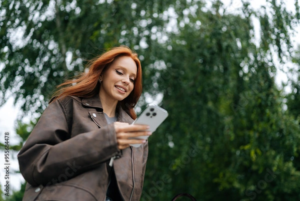 Fototapeta Low-angle view of young woman with long red hair joyfully texting on mobile phone while strolling through city street on summer day. Cheerful redhead female happily messaging on smartphone outdoors.