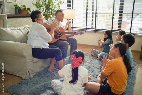 Fototapeta Elderly couple playing guitar and clapping along with a children seated on the floor in a cozy living room. The scene captures the joy of intergenerational bonding through music family atmosphere