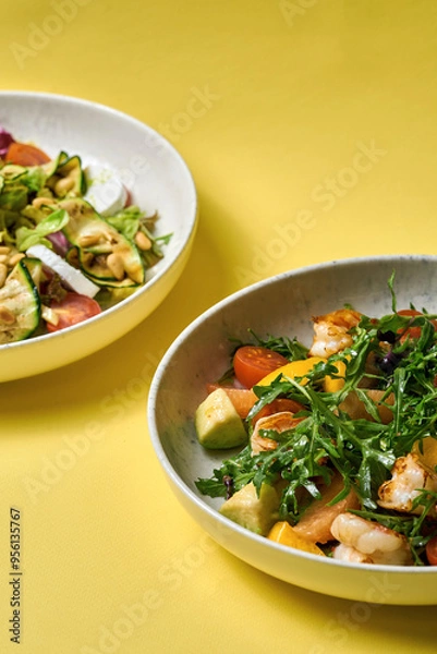 Fototapeta A fresh and vibrant salad featuring grilled shrimp, arugula, cherry tomatoes, avocado, and citrus segments, served in a modern bowl against a bright yellow background.