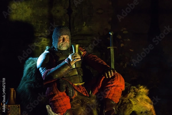 Obraz Old medieval King in armor with dish is sitting on furs near the