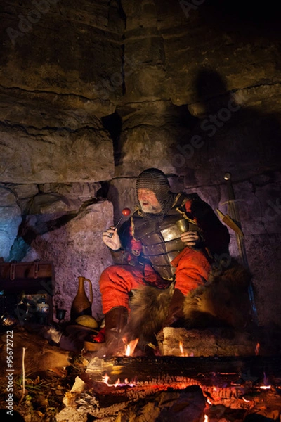 Obraz Old medieval King in armor with dish is sitting on furs near the