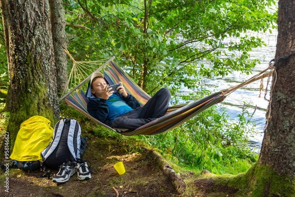 Obraz A teenage boy is relaxing in a hammock on the shore of a forest lake and playing with a smartphone.