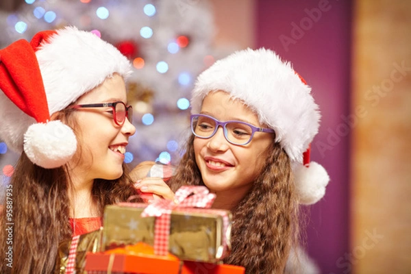 Obraz Happy sisters holding gifts in front of christmas tree