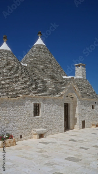 Fototapeta Traditional trulli house in alberobello, italy with conical roofs, white stone walls, and a vivid blue sky in the background.