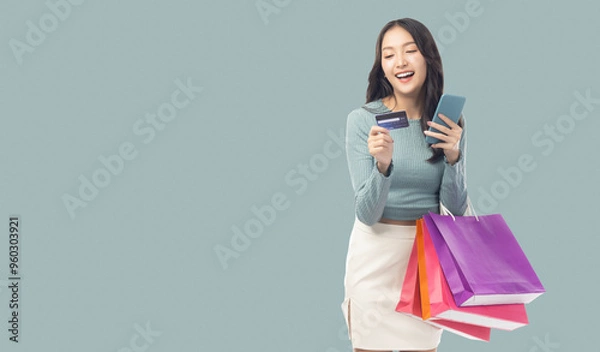 Fototapeta Young asian lady using mobile phone holding credit card carry shopping bags isolated over pastel green background and copy space Smiling young girl purchasing online through phone using credit card