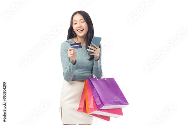 Fototapeta Young asian lady using mobile phone holding credit card carry shopping bags isolated over pastel green background and copy space Smiling young girl purchasing online through phone using credit card