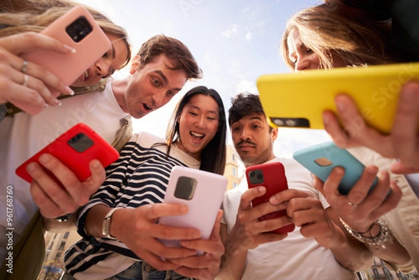 Fototapeta Diverse group young people in circle staring in surprise at cell phone screen together outdoors. Amazed generation z student friends using colorful mobiles enjoying interesting social media content 