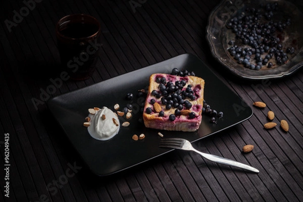 Obraz A rustic dessert featuring a slice of blueberry cake on a black plate, served with a dollop of whipped cream and almonds. Fresh blueberries and almonds scattered around enhance the dark, cozy mood