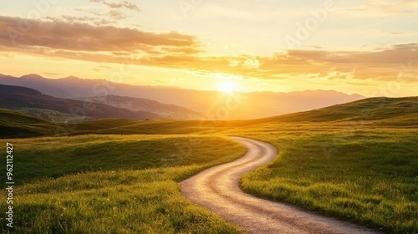 Fototapeta A serene landscape with a winding road through green fields, illuminated by a beautiful sunset over distant mountains.