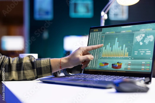 Obraz An individual pointing at a screen that shows numbers and financial charts related to stock trading. At a desk, digital laptop is displaying graphs and values for forex trends.
