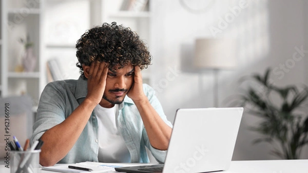 Obraz A young Indian male businessman is sitting at a desk trying to work, checking schedules and getting frustrated that something is not working out. Headache, fatigue, burnout.