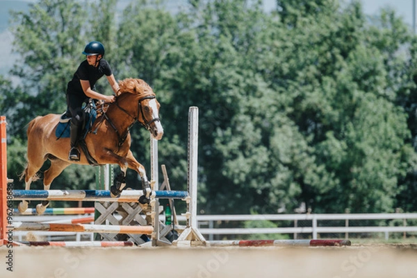 Fototapeta Equestrian rider skillfully jumps a brown horse over an obstacle during an outdoor training session. The focus is on the synergy between the horse and rider.