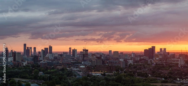 Fototapeta A panoramic view of Manchester city skyline at sunset, with dramatic clouds and a warm color palette.
