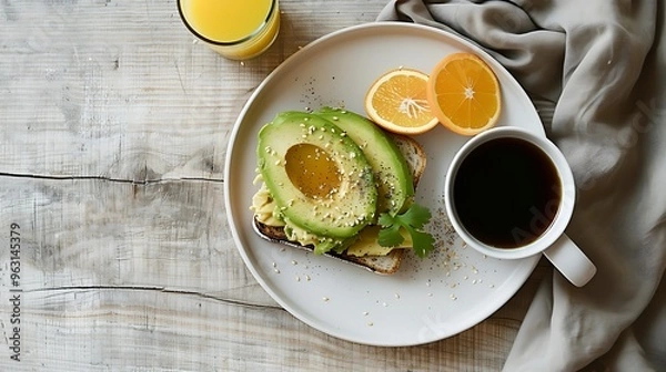 Fototapeta Morning meal featuring avocado toast on a white plate with a mug of coffee and a glass of orange juice set on a light wooden table with a linen napkin for a fresh start