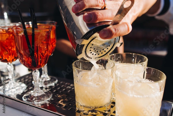 Fototapeta Bartender working, beautiful row line of different coloured liquor alcohol cocktails on a open air party in a bar, and others on catering banquet on event, barman mixing drinks and beverages in club