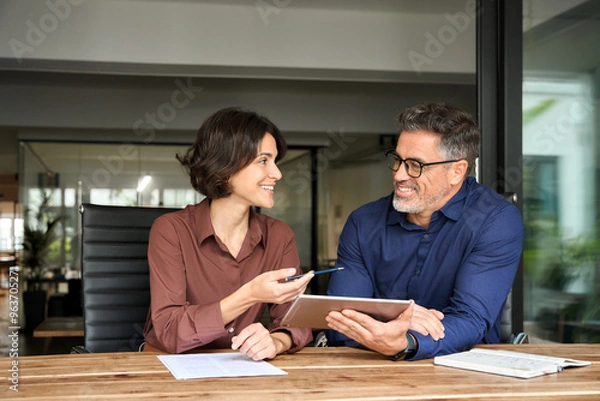 Obraz Two happy busy business colleagues having conversation at corporate meeting. Business team people having discussion planning sales financial strategy in company using tablet working together in office