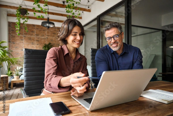 Fototapeta Business team of two executives working together using laptop in office. Happy young business woman manager showing project presentation to mature man client, investor or partner at work meeting.