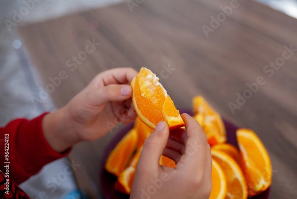 Fototapeta a close-up of a child’s hands holding a slice of orange, with a plate of orange slices on a wooden surface in the background.
