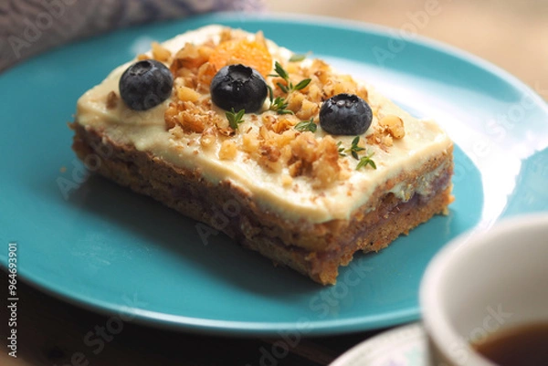Fototapeta A vegan dessert featuring moist cake topped with creamy frosting, walnuts, fresh blueberries, and thyme. A perfect plant-based treat for a relaxing coffee break.