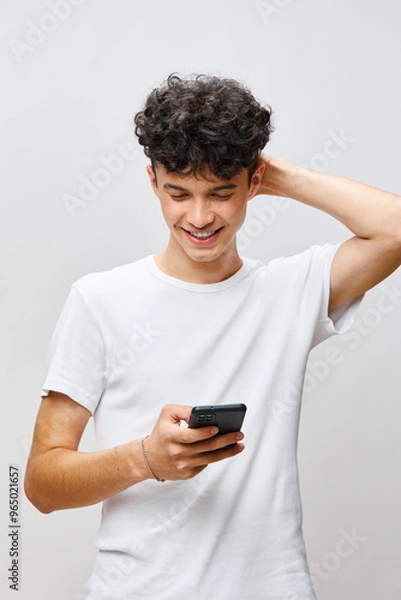 Fototapeta Young man using smartphone, smiling happily, wearing a plain white t shirt, standing against a light grey background, showcasing a sense of joy and connectivity