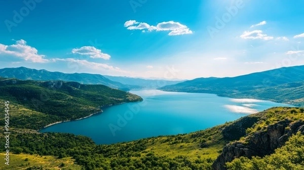 Obraz A breathtaking panoramic view showcasing a crystal clear lake nestled amidst lush green mountains under a vibrant blue sky. The scene evokes a sense of tranquility, vastness, and natural beauty.