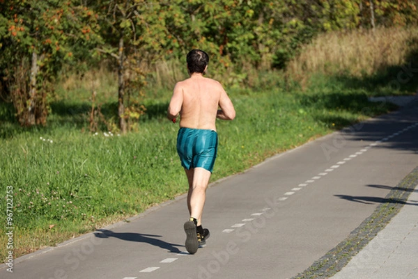 Fototapeta Man in shorts running on sidewalk along the city street, rear view. Runner in park, workout in autumn season