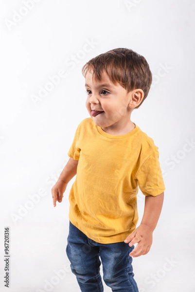 Obraz Portrait of a two year old boy looking shy sticking out his tongue while looking away on white background