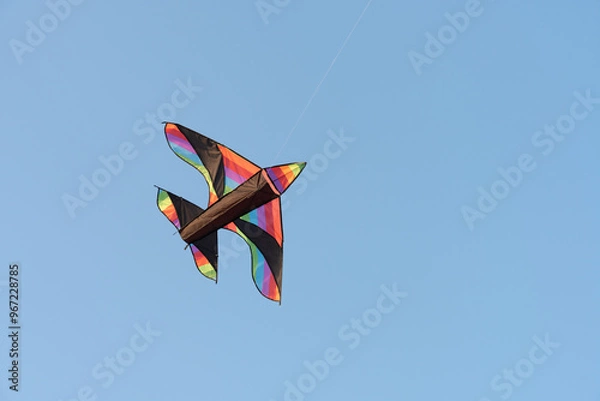 Fototapeta View to the flying kite in the blue sky. The kite has shape of bird or airplane and colourful body. Great experience for kids and adults. 