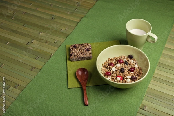 Obraz A healthy breakfast setup featuring a bowl of granola with dried fruits and white chocolate chips, a granola bar, and a cup of milk. The scene is arranged on a green placemat.