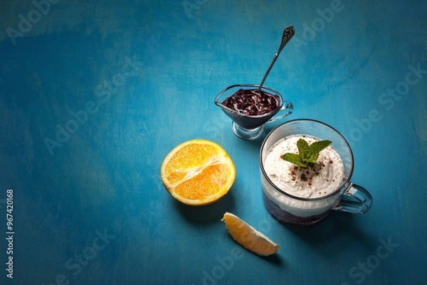 Fototapeta A delightful dessert arrangement with a glass of whipped cream layered with blueberry compote, garnished with mint, served alongside a halved orange and a small dish of blueberry jam on a blue table.