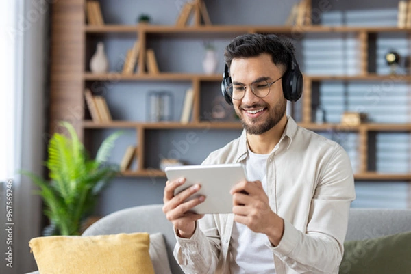 Obraz A young Indian man wearing headphones is sitting on the sofa at home, holding a tablet and looking at the screen with a smile