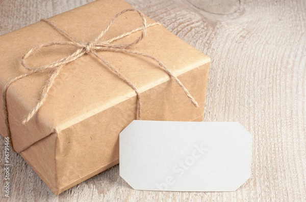 Obraz Gift box into brown paper tied by twine with blank white card on white wooden table with space for text