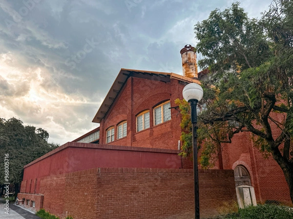 Fototapeta Vintage Industrial Red Brick Building with Rusted Chimney and Lamppost Under a Moody Sky in Monterrey
