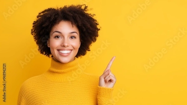 Obraz A cheerful woman wearing a yellow sweater, smiling and pointing at something on a colorful background