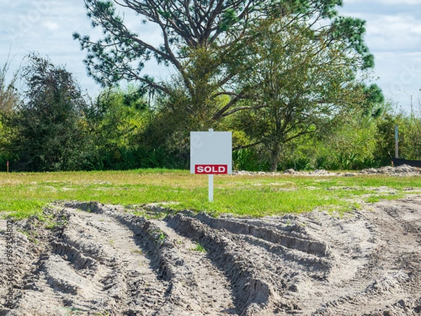 Obraz Street view of "Sold" sign on a vacant lot with recent tire ruts in sand in a suburban residential development on a sunny afternoon in southwest Florida. Motifs of real estate, commitment, the future.