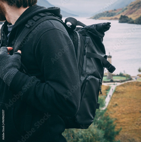 Obraz Crop of a male hiker and his backpack as he stands near some hills at Glenfinnan Monument in the Highlands, Scotland. He wears a non-branded camera backpack, black hoodie and fingerless gloves.