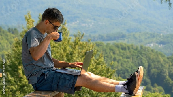 Fototapeta Middle aged man works with laptop and drinks coffee in nature on background of mountains