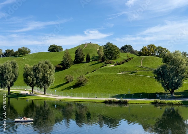 Fototapeta People enjoying Munich's Olympic Park grounds where there is a beautiful lake where boats can be rented which is surrounded by a picturesque grassy hill with a sweeping view.